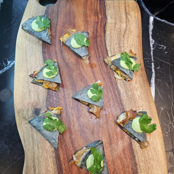 Seven triangular blue corn quesadillas with filling, green sauce, and cilantro on a wooden board.