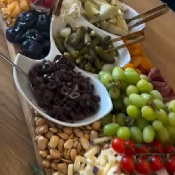 A charcuterie board with olives, pickles, grapes, tomatoes, nuts, and cheeses.