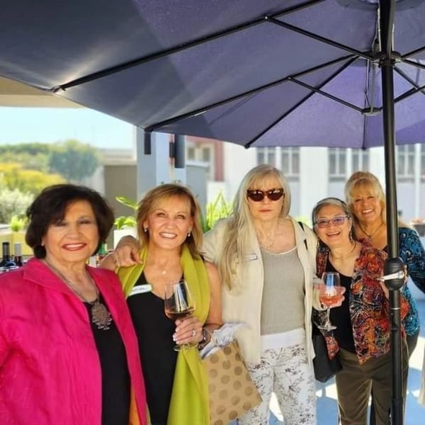 Five smiling women pose together outdoors under a dark umbrella, holding wine glasses.