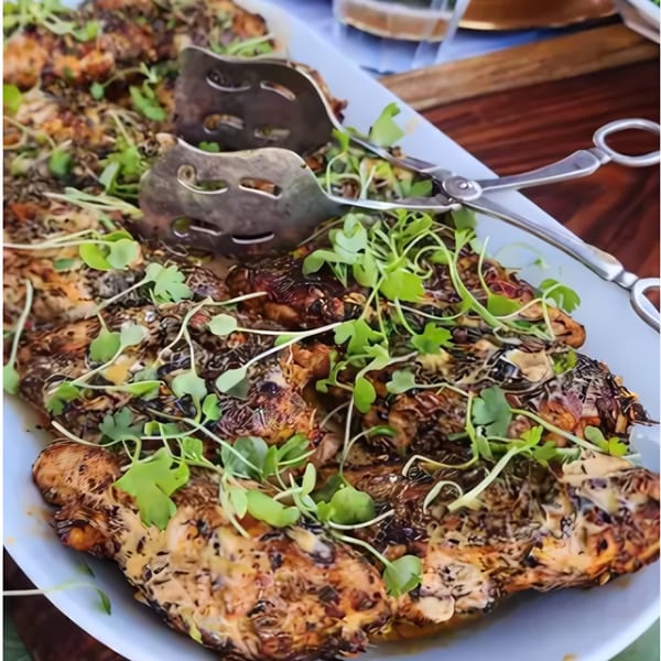Grilled chicken pieces with a creamy sauce and fresh microgreens on a white platter with serving tongs.