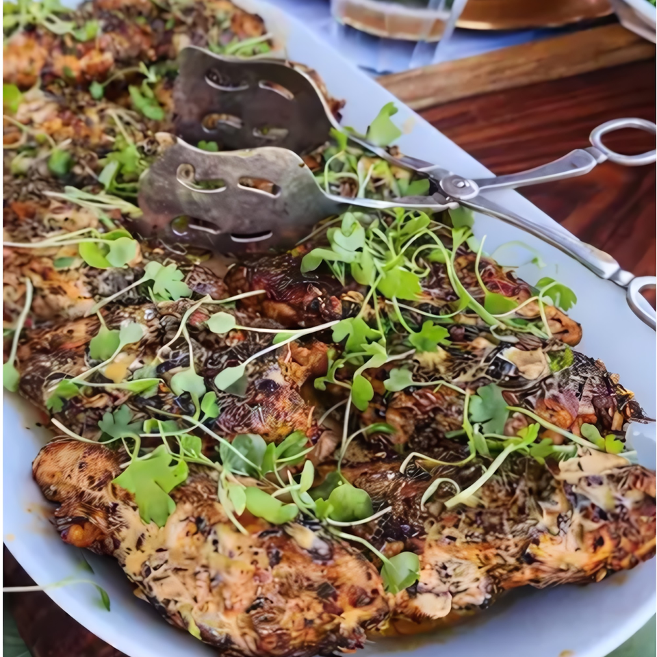 Grilled chicken pieces with a creamy sauce and fresh microgreens on a white platter with serving tongs.