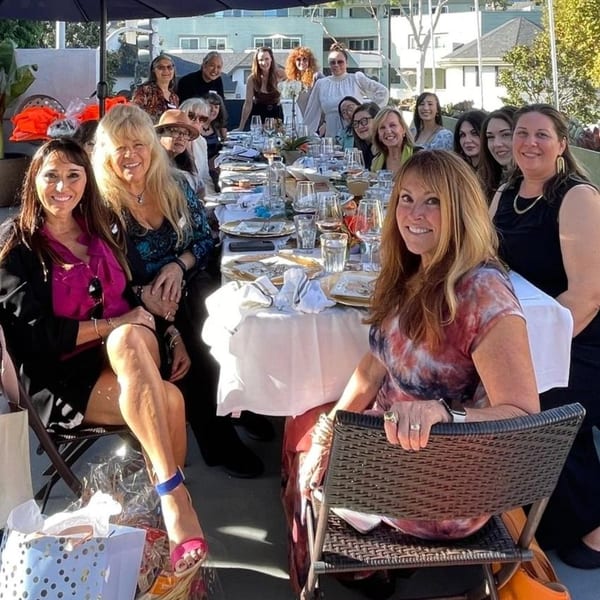 A group of women smiling and posing for a photo around a long dining table outdoors.