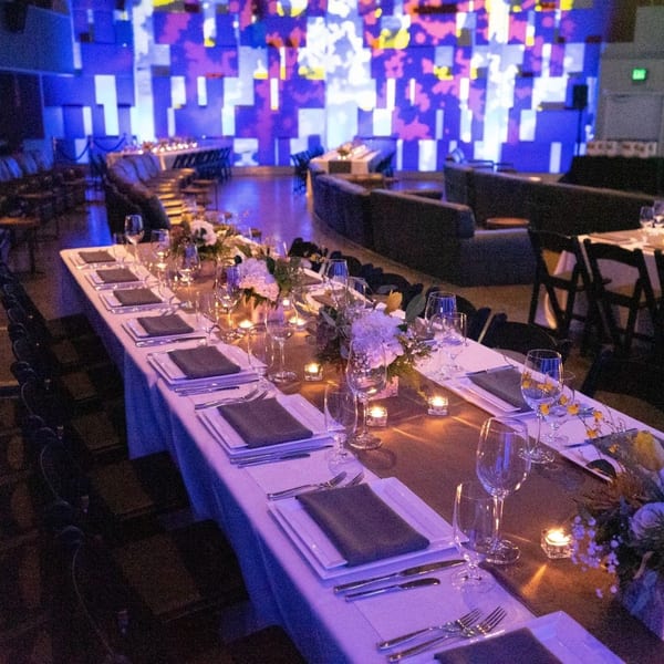 Long dining tables set with white plates, dark napkins, and floral centerpieces under blue lighting.