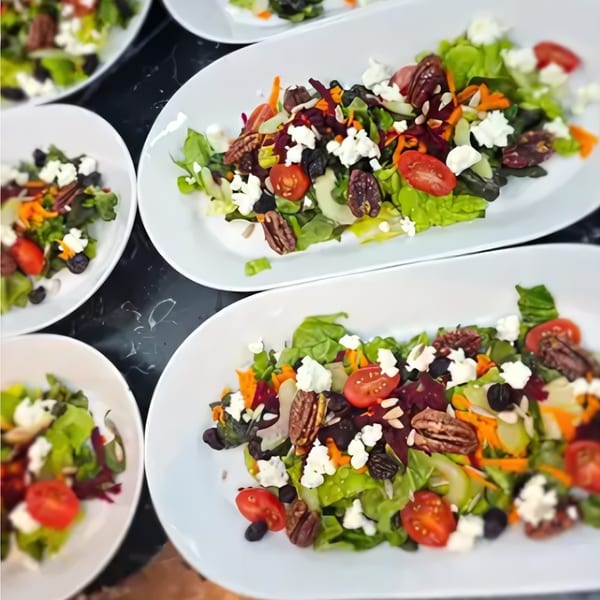 Multiple plates of fresh green salad with tomatoes, feta, pecans, and olives on a dark surface.