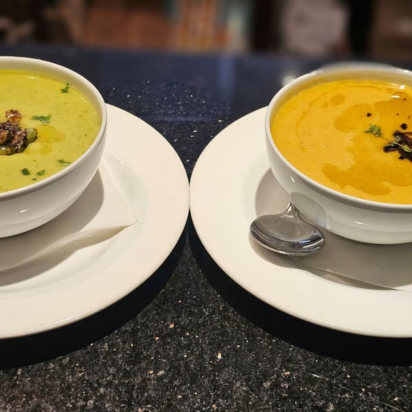 Two bowls of soup, one green and one orange, on white plates with spoons on a dark speckled counter.