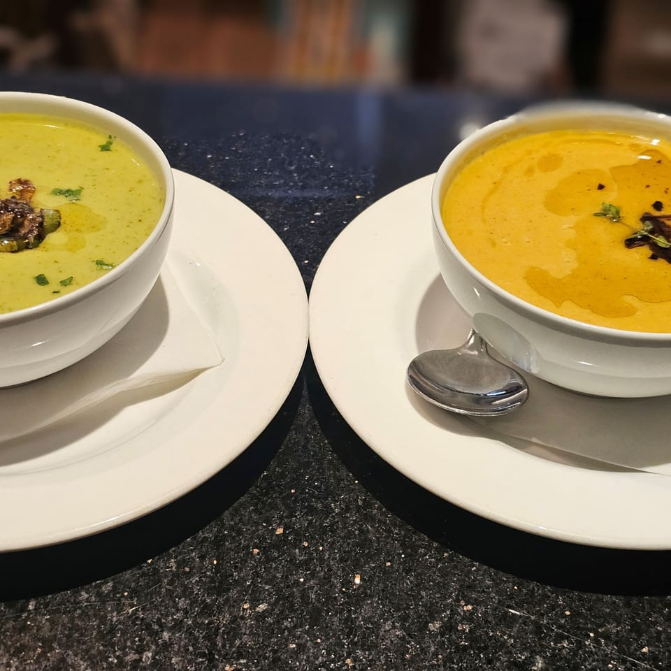 Two bowls of soup, one green and one orange, on white plates with spoons on a dark speckled counter.