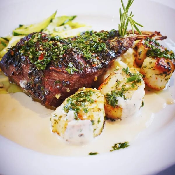 Grilled steak with herbs, seared scallops, and mashed potatoes in a creamy sauce on a white plate.