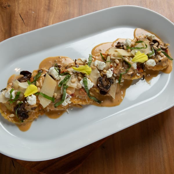 Ravioli with creamy sauce, mushrooms, pine nuts, cheese, basil, and yellow edible flowers on a white plate.
