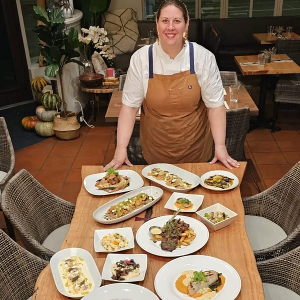 Chef Lindsay smiles behind a table laden with various American dishes at Nirvana restaurant.